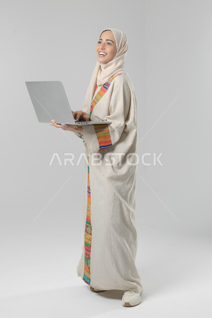 Saudi women's jobs and professions, business management using technology and modern devices, portrait of a smiling Saudi Arabian Gulf woman wearing an abaya and hijab holding a laptop, smart and secure e-marketing, full body image, white background
