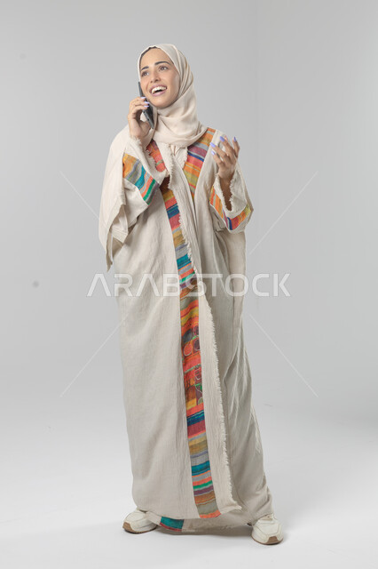 التحدث عبر الهاتف المحمول، قضاء اوقات ممتعة بتبادل الاخبار والأحاديث، بورتريه لامرأة عربية خليجية سعودية محجبة ترتدي العباءة تقوم بإجرء مكالمة هاتفية، محادثات ودردشات مع الاصدقاء، صورة الجسم بالكامل، خلفية بيضاء