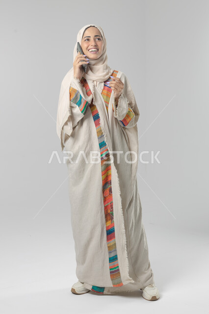 التحدث عبر الهاتف المحمول، قضاء اوقات ممتعة بتبادل الاخبار والأحاديث، بورتريه لامرأة عربية خليجية سعودية محجبة ترتدي العباءة تقوم بإجرء مكالمة هاتفية، محادثات ودردشات مع الاصدقاء، صورة الجسم بالكامل، خلفية بيضاء