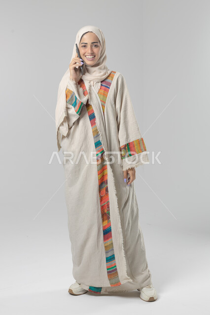 التحدث عبر الهاتف المحمول، قضاء اوقات ممتعة بتبادل الاخبار والأحاديث، بورتريه لامرأة عربية خليجية سعودية محجبة ترتدي العباءة تقوم بإجرء مكالمة هاتفية، محادثات ودردشات مع الاصدقاء، صورة الجسم بالكامل، خلفية بيضاء