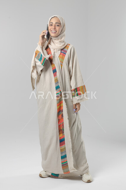 التحدث عبر الهاتف المحمول، قضاء اوقات ممتعة بتبادل الاخبار والأحاديث، بورتريه لامرأة عربية خليجية سعودية محجبة ترتدي العباءة تقوم بإجرء مكالمة هاتفية، محادثات ودردشات مع الاصدقاء، صورة الجسم بالكامل، خلفية بيضاء