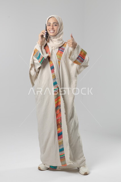 التحدث عبر الهاتف المحمول، قضاء اوقات ممتعة بتبادل الاخبار والأحاديث، بورتريه لامرأة عربية خليجية سعودية محجبة ترتدي العباءة تقوم بإجرء مكالمة هاتفية، محادثات ودردشات مع الاصدقاء، صورة الجسم بالكامل، خلفية بيضاء