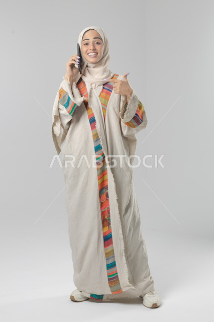 التحدث عبر الهاتف المحمول، قضاء اوقات ممتعة بتبادل الاخبار والأحاديث، بورتريه لامرأة عربية خليجية سعودية محجبة ترتدي العباءة تقوم بإجرء مكالمة هاتفية، محادثات ودردشات مع الاصدقاء، صورة الجسم بالكامل، خلفية بيضاء