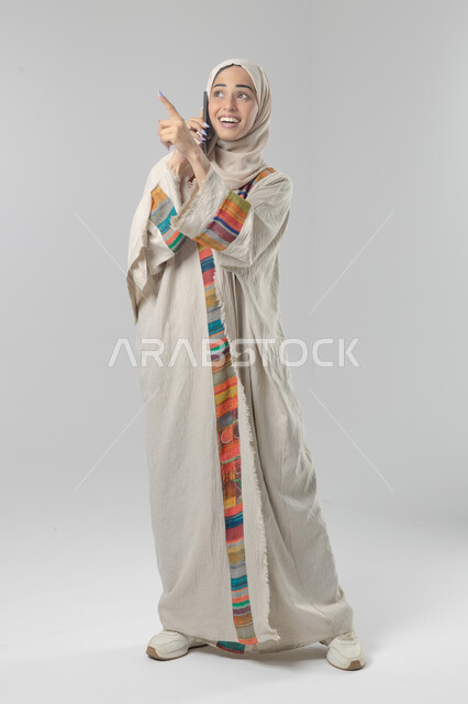 التحدث عبر الهاتف المحمول، قضاء اوقات ممتعة بتبادل الاخبار والأحاديث، بورتريه لامرأة عربية خليجية سعودية محجبة ترتدي العباءة تقوم بإجرء مكالمة هاتفية، محادثات ودردشات مع الاصدقاء، صورة الجسم بالكامل، خلفية بيضاء
