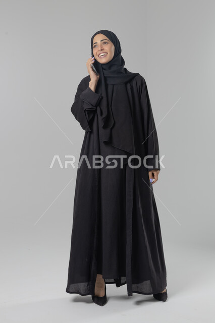 قضاء اوقات ممتعة بتبادل الاخبار والأحاديث، التحدث عبر الهاتف المحمول، بورتريه لامرأة عربية خليجية سعودية محجبة ترتدي العباءة تقوم بإجرء مكالمة هاتفية، محادثات ودردشات مع الاصدقاء، صورة الجسم بالكامل، خلفية بيضاء