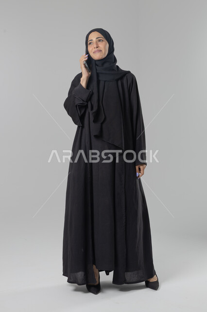 قضاء اوقات ممتعة بتبادل الاخبار والأحاديث، التحدث عبر الهاتف المحمول، بورتريه لامرأة عربية خليجية سعودية محجبة ترتدي العباءة تقوم بإجرء مكالمة هاتفية، محادثات ودردشات مع الاصدقاء، صورة الجسم بالكامل، خلفية بيضاء