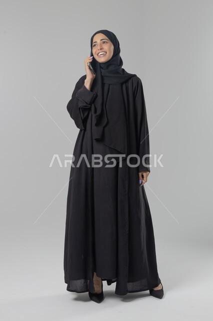 قضاء اوقات ممتعة بتبادل الاخبار والأحاديث، التحدث عبر الهاتف المحمول، بورتريه لامرأة عربية خليجية سعودية محجبة ترتدي العباءة تقوم بإجرء مكالمة هاتفية، محادثات ودردشات مع الاصدقاء، صورة الجسم بالكامل، خلفية بيضاء