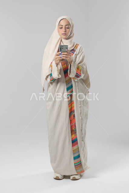  تعابير وايماءات الاندماج والسرور، استخدام التقنية الحديثه المتطورة في الحياة العملية، بورتريه لإمرأة عربية خليجية سعودية ترتدي الحجاب والعباءة تقوم بالعمل على الهاتف المحمول، صورة الجسم بالطول الكامل، خلفية بيضاء 