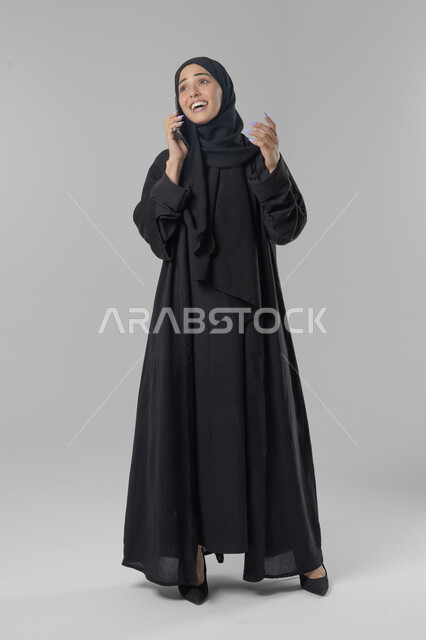 قضاء اوقات ممتعة بتبادل الاخبار والأحاديث، التحدث عبر الهاتف المحمول، بورتريه لامرأة عربية خليجية سعودية محجبة ترتدي العباءة تقوم بإجرء مكالمة هاتفية، محادثات ودردشات مع الاصدقاء، صورة الجسم بالكامل، خلفية بيضاء