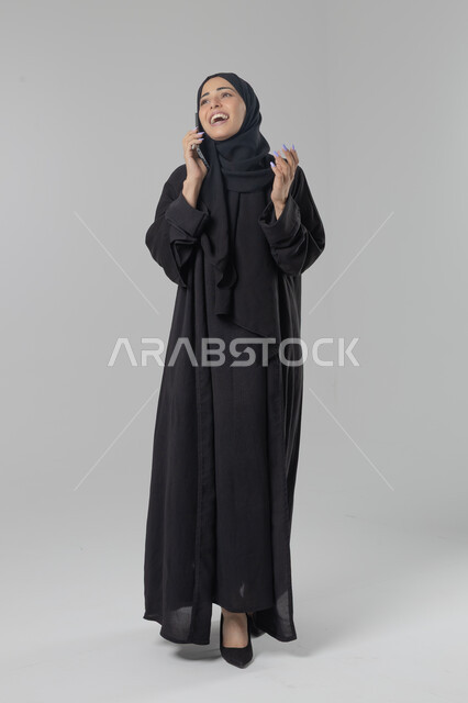 Having a good time exchanging news and conversations, talking on mobile phone, portrait of a veiled Saudi Arabian Gulf woman wearing an abaya making a phone call, conversations and chats with friends, full body image, white background