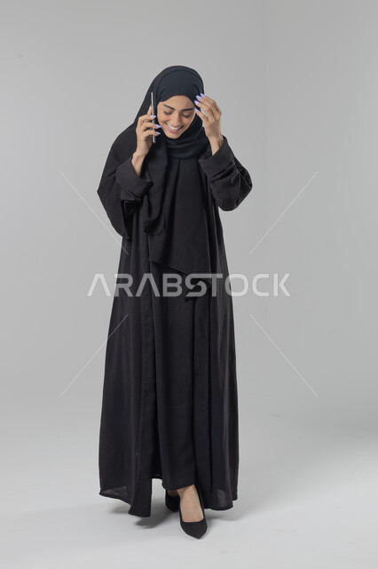 قضاء اوقات ممتعة بتبادل الاخبار والأحاديث، التحدث عبر الهاتف المحمول، بورتريه لامرأة عربية خليجية سعودية محجبة ترتدي العباءة تقوم بإجرء مكالمة هاتفية، محادثات ودردشات مع الاصدقاء، صورة الجسم بالكامل، خلفية بيضاء