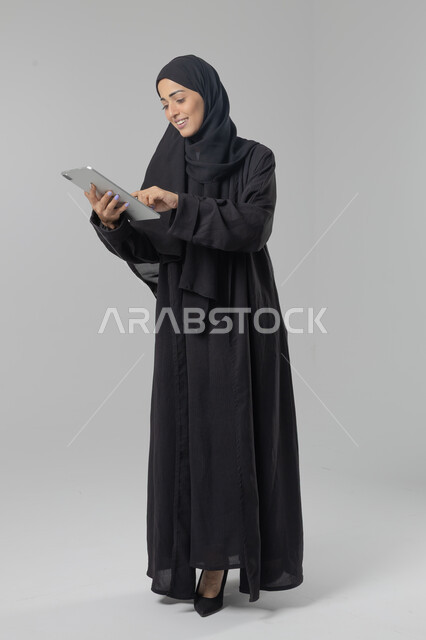 Using modern advanced technology in practical life via tablet, expressions and gestures of integration, portrait of a young Saudi Arabian Gulf woman wearing hijab and abaya working on a tablet, full body image, white background