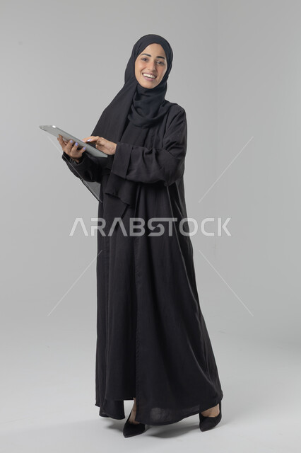 Using modern advanced technology in practical life via tablet, expressions and gestures of integration, portrait of a young Saudi Arabian Gulf woman wearing hijab and abaya working on a tablet, full body image, white background