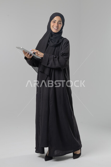  استخدام التقنية الحديثه المتطورة في الحياة العملية عبر الجهاز اللوحي، تعابير وايماءات الاندماج، بورتريه لشابة عربية خليجية سعودية ترتدي الحجاب والعباءة تقوم بالعمل على جهازالتابلت، صورة الجسم بالطول الكامل، خلفية بيضاء 