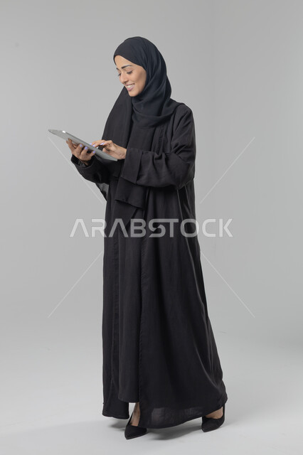  استخدام التقنية الحديثه المتطورة في الحياة العملية عبر الجهاز اللوحي، تعابير وايماءات الاندماج، بورتريه لشابة عربية خليجية سعودية ترتدي الحجاب والعباءة تقوم بالعمل على جهازالتابلت، صورة الجسم بالطول الكامل، خلفية بيضاء 
