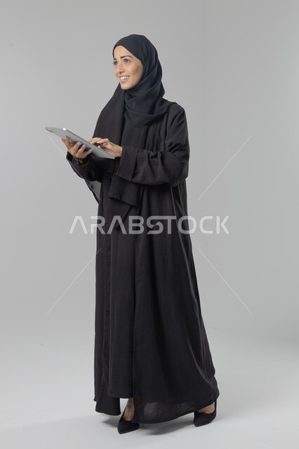 Using modern advanced technology in practical life via tablet, expressions and gestures of integration, portrait of a young Saudi Arabian Gulf woman wearing hijab and abaya working on a tablet, full body image, white background