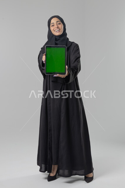 عرض شاشة كروما فارغة عبر الجهاز اللوحي، استخدام التقنية الحديثه المتطورة في الحياة العملية، تعابير وايماءات الاندماج، بورتريه لشابة عربية خليجية سعودية ترتدي الحجاب والعباءة تقوم بالعمل على جهازالتابلت، صورة الجسم بالطول الكامل، خلفية بيضاء 