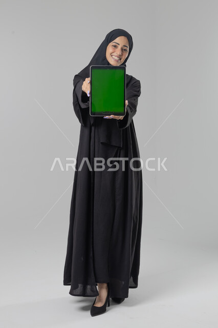 Blank screen display on tablet, use of modern advanced technology in practical life, expressions and gestures of integration, portrait of a young Saudi Arabian Gulf woman wearing hijab and abaya working on a tablet, full body image, white background