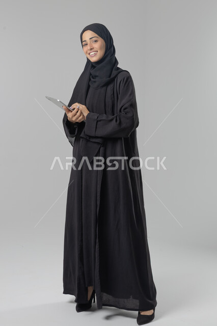  استخدام التقنية الحديثه المتطورة في الحياة العملية عبر الجهاز اللوحي، تعابير وايماءات الاندماج، بورتريه لشابة عربية خليجية سعودية ترتدي الحجاب والعباءة تقوم بالعمل على جهازالتابلت، صورة الجسم بالطول الكامل، خلفية بيضاء 