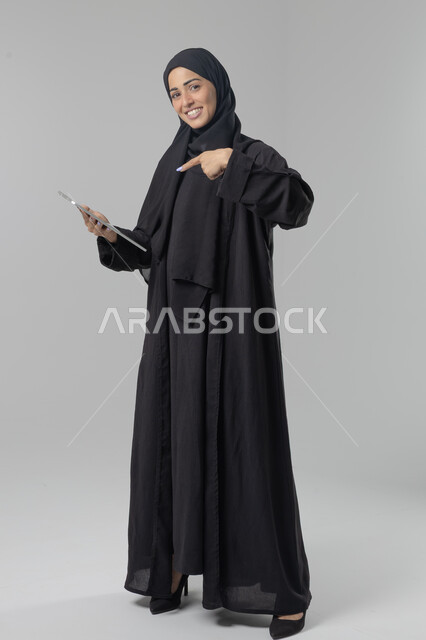 Using modern advanced technology in practical life via tablet, expressions and gestures of integration, portrait of a young Saudi Arabian Gulf woman wearing hijab and abaya working on a tablet, full body image, white background
