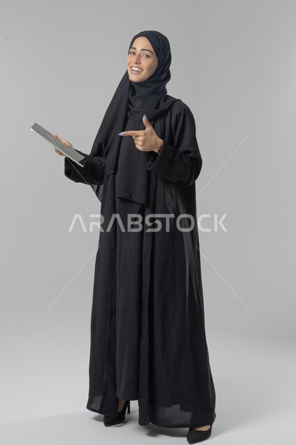 Using modern advanced technology in practical life via tablet, expressions and gestures of integration, portrait of a young Saudi Arabian Gulf woman wearing hijab and abaya working on a tablet, full body image, white background
