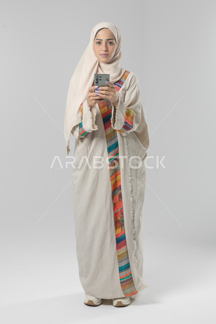  تعابير وايماءات الاندماج والسرور، استخدام التقنية الحديثه المتطورة في الحياة العملية، بورتريه لإمرأة عربية خليجية سعودية ترتدي الحجاب والعباءة تقوم بالعمل على الهاتف المحمول، صورة الجسم بالطول الكامل، خلفية بيضاء 