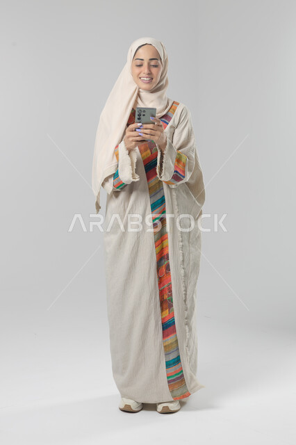  تعابير وايماءات الاندماج والسرور، استخدام التقنية الحديثه المتطورة في الحياة العملية، بورتريه لإمرأة عربية خليجية سعودية ترتدي الحجاب والعباءة تقوم بالعمل على الهاتف المحمول، صورة الجسم بالطول الكامل، خلفية بيضاء 