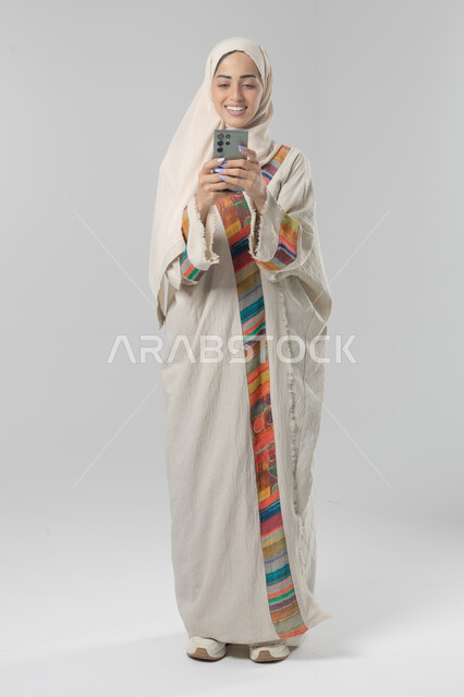  تعابير وايماءات الاندماج والسرور، استخدام التقنية الحديثه المتطورة في الحياة العملية، بورتريه لإمرأة عربية خليجية سعودية ترتدي الحجاب والعباءة تقوم بالعمل على الهاتف المحمول، صورة الجسم بالطول الكامل، خلفية بيضاء 