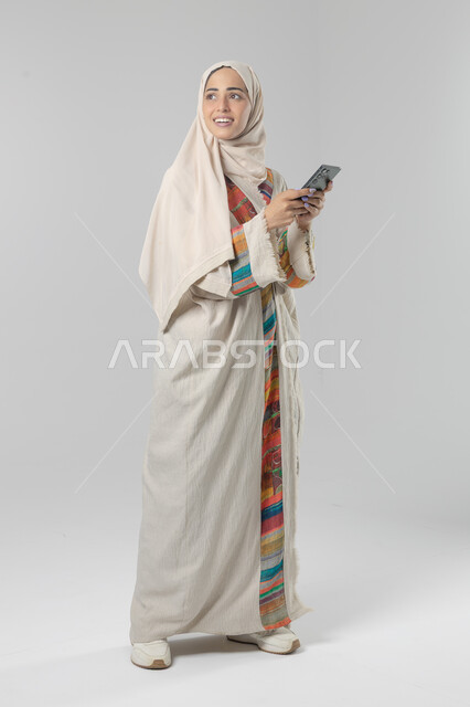 تعابير وايماءات الاندماج والسرور، استخدام التقنية الحديثه المتطورة في الحياة العملية، بورتريه لإمرأة عربية خليجية سعودية ترتدي الحجاب والعباءة تقوم بالعمل على الهاتف المحمول، صورة الجسم بالطول الكامل، خلفية بيضاء 