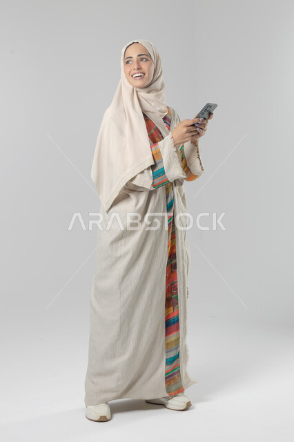  تعابير وايماءات الاندماج والسرور، استخدام التقنية الحديثه المتطورة في الحياة العملية، بورتريه لإمرأة عربية خليجية سعودية ترتدي الحجاب والعباءة تقوم بالعمل على الهاتف المحمول، صورة الجسم بالطول الكامل، خلفية بيضاء 