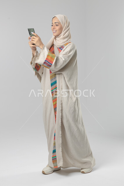 Expressions and gestures of integration and pleasure, use of modern advanced technology in practical life, portrait of a Saudi Arabian Gulf woman wearing a hijab and abaya working on a mobile phone, full body image, white background