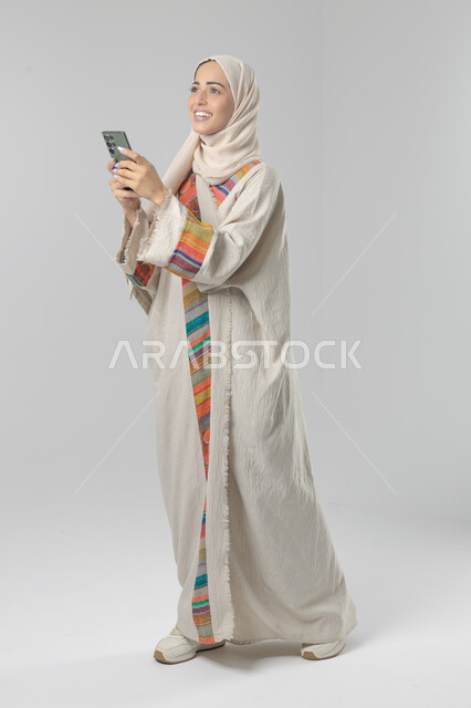  تعابير وايماءات الاندماج والسرور، استخدام التقنية الحديثه المتطورة في الحياة العملية، بورتريه لإمرأة عربية خليجية سعودية ترتدي الحجاب والعباءة تقوم بالعمل على الهاتف المحمول، صورة الجسم بالطول الكامل، خلفية بيضاء 