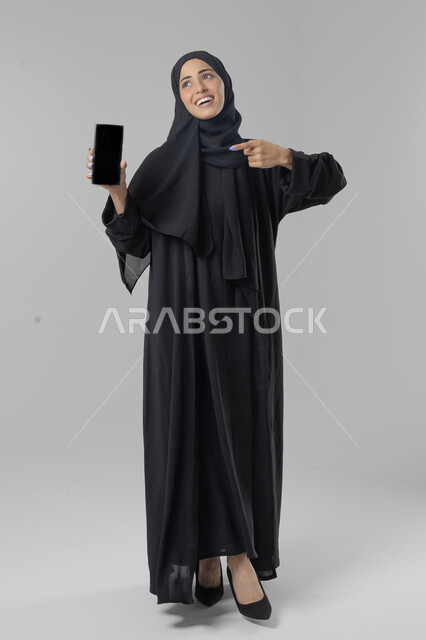 Empty black screen display, expressions and gestures of integration and pleasure, use of modern advanced technology in practical life, portrait of a Saudi Arabian Gulf woman wearing hijab and abaya working on a mobile phone, full body image, white background
