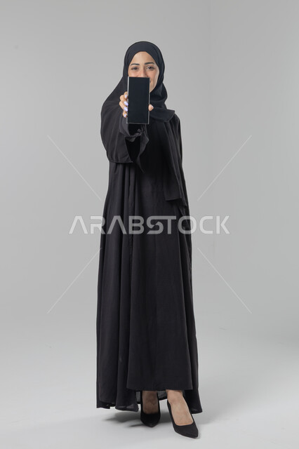 عرض شاشة سوداء فارغة، تعابير وايماءات الاندماج والسرور، استخدام التقنية الحديثه المتطورة في الحياة العملية، بورتريه لإمرأة عربية خليجية سعودية ترتدي الحجاب والعباءة تقوم بالعمل على الهاتف المحمول، صورة الجسم بالطول الكامل، خلفية بيضاء 