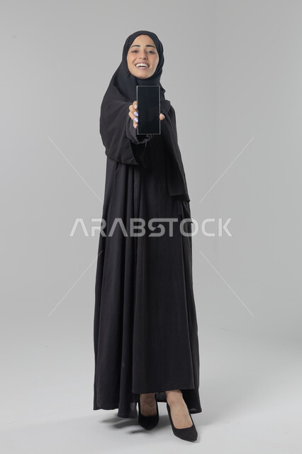 عرض شاشة سوداء فارغة، تعابير وايماءات الاندماج والسرور، استخدام التقنية الحديثه المتطورة في الحياة العملية، بورتريه لإمرأة عربية خليجية سعودية ترتدي الحجاب والعباءة تقوم بالعمل على الهاتف المحمول، صورة الجسم بالطول الكامل، خلفية بيضاء 