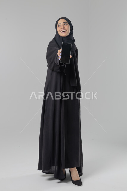 عرض شاشة سوداء فارغة، تعابير وايماءات الاندماج والسرور، استخدام التقنية الحديثه المتطورة في الحياة العملية، بورتريه لإمرأة عربية خليجية سعودية ترتدي الحجاب والعباءة تقوم بالعمل على الهاتف المحمول، صورة الجسم بالطول الكامل، خلفية بيضاء 