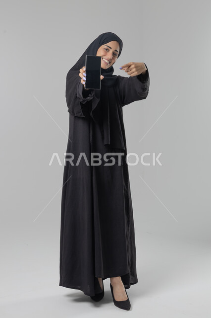 عرض شاشة سوداء فارغة، تعابير وايماءات الاندماج والسرور، استخدام التقنية الحديثه المتطورة في الحياة العملية، بورتريه لإمرأة عربية خليجية سعودية ترتدي الحجاب والعباءة تقوم بالعمل على الهاتف المحمول، صورة الجسم بالطول الكامل، خلفية بيضاء 