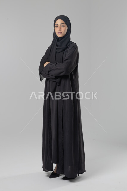 Looking at the camera with gestures of pleasure and self-confidence, the concept of elegance and interest in external appearance, a portrait of a Saudi Arabian Gulf woman wearing the hijab and traditional abaya standing with crossed hands, full-length body image, white background