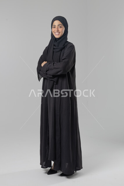 النظر إلى الكاميرا بإيماءات السرور والثقة بالنفس، مفهوم الاناقه و الاهتمام بالمظهر الخارجي، بورتريه لإمرأة عربية خليجية سعودية ترتدي الحجاب والعباءة التقليدية تقف بأيدي متقاطعه، صورة الجسم بالطول الكامل، خلفية بيضاء