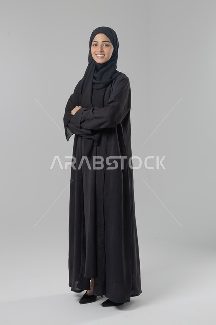 النظر إلى الكاميرا بإيماءات السرور والثقة بالنفس، مفهوم الاناقه و الاهتمام بالمظهر الخارجي، بورتريه لإمرأة عربية خليجية سعودية ترتدي الحجاب والعباءة التقليدية تقف بأيدي متقاطعه، صورة الجسم بالطول الكامل، خلفية بيضاء