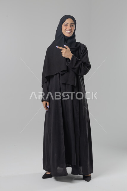  التوضيح والدلالة لمكان محدد, الإشارة إلى عنصر ما, بورتريه لامرأة عربية خليجية سعودية محجبة ترتدي العباءة تشير بيديها على الجانب، تحديد موقع شيء معين, صورة الجسم بالطول الكامل، خلفية بيضاء