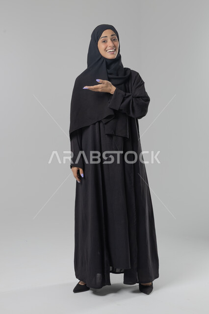  التوضيح والدلالة لمكان محدد, الإشارة إلى عنصر ما, بورتريه لامرأة عربية خليجية سعودية محجبة ترتدي العباءة تشير بيديها على الجانب، تحديد موقع شيء معين, صورة الجسم بالطول الكامل، خلفية بيضاء