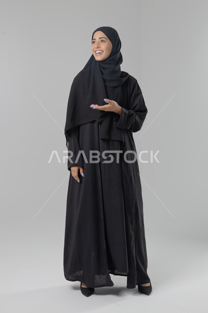  التوضيح والدلالة لمكان محدد, الإشارة إلى عنصر ما, بورتريه لامرأة عربية خليجية سعودية محجبة ترتدي العباءة تشير بيديها على الجانب، تحديد موقع شيء معين, صورة الجسم بالطول الكامل، خلفية بيضاء