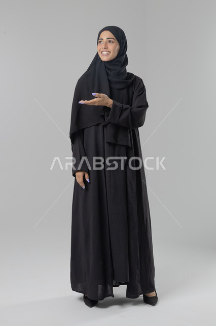  التوضيح والدلالة لمكان محدد, الإشارة إلى عنصر ما, بورتريه لامرأة عربية خليجية سعودية محجبة ترتدي العباءة تشير بيديها على الجانب، تحديد موقع شيء معين, صورة الجسم بالطول الكامل، خلفية بيضاء
