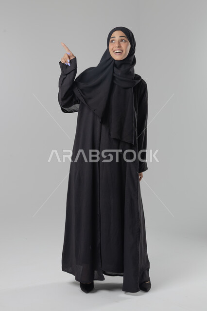  التوضيح والدلالة لمكان محدد, الإشارة إلى عنصر ما, بورتريه لامرأة عربية خليجية سعودية محجبة ترتدي العباءة تشير بيديها على الجانب، تحديد موقع شيء معين, صورة الجسم بالطول الكامل، خلفية بيضاء