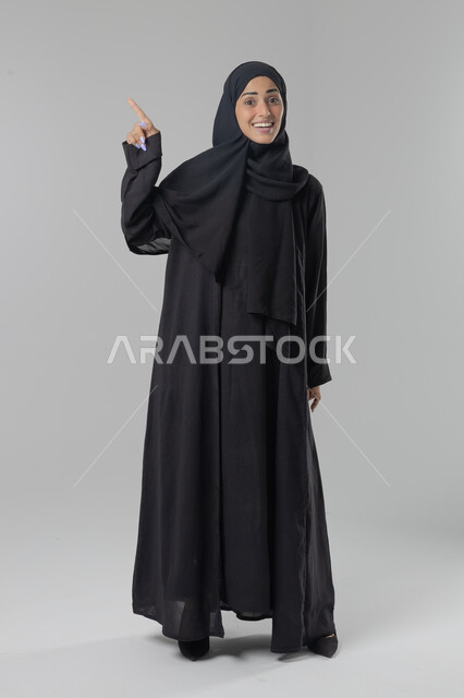  التوضيح والدلالة لمكان محدد, الإشارة إلى عنصر ما, بورتريه لامرأة عربية خليجية سعودية محجبة ترتدي العباءة تشير بيديها على الجانب، تحديد موقع شيء معين, صورة الجسم بالطول الكامل، خلفية بيضاء