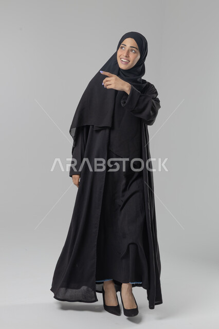  التوضيح والدلالة لمكان محدد, الإشارة إلى عنصر ما, بورتريه لامرأة عربية خليجية سعودية محجبة ترتدي العباءة تشير بيديها على الجانب، تحديد موقع شيء معين, صورة الجسم بالطول الكامل، خلفية بيضاء