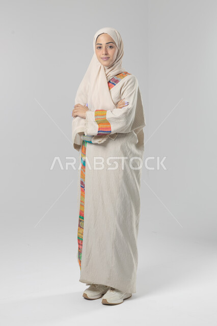 Looking at the camera with gestures of pleasure and self-confidence, the concept of elegance and interest in external appearance, a portrait of a Saudi Arabian Gulf woman wearing the hijab and traditional abaya standing with crossed hands, full-length body image, white background