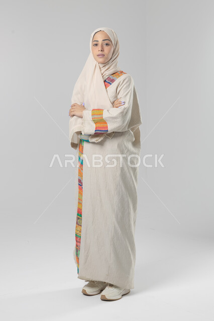 Looking at the camera with gestures of pleasure and self-confidence, the concept of elegance and interest in external appearance, a portrait of a Saudi Arabian Gulf woman wearing the hijab and traditional abaya standing with crossed hands, full-length body image, white background