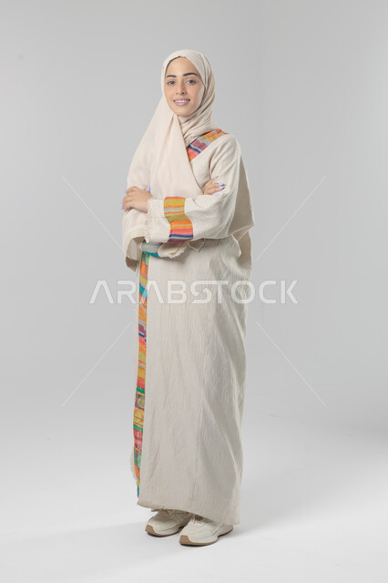 النظر إلى الكاميرا بإيماءات السرور والثقة بالنفس، مفهوم الاناقه و الاهتمام بالمظهر الخارجي، بورتريه لإمرأة عربية خليجية سعودية ترتدي الحجاب والعباءة التقليدية تقف بأيدي متقاطعه، صورة الجسم بالطول الكامل، خلفية بيضاء